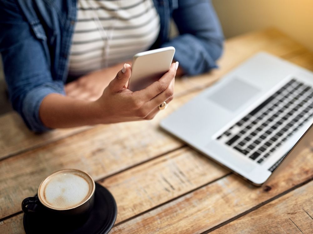 Mulher segurando o celular enquanto trabalha no laptop, refletindo sobre o que fazer para atrair clientes usando tecnologia e estratégias de marketing digital.