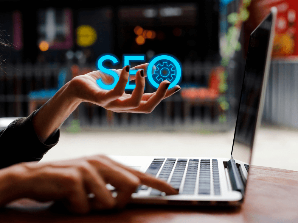 Pessoa utilizando notebook enquanto o termo SEO aparece em destaque, representando estratégias de otimização para marketing digital.