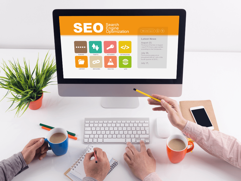 Computador exibindo uma página sobre SEO enquanto duas pessoas trabalham, representando a otimização de sites e análise digital.