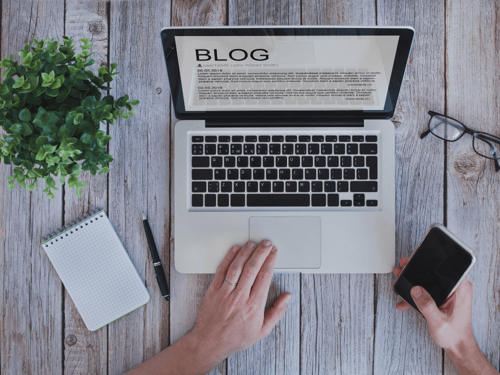 Pessoa trabalha em notebook com uma página de blog aberta, acompanhada de caderno e celular sobre uma mesa de madeira. Tudo para explicar como fazer um blog de sucesso.