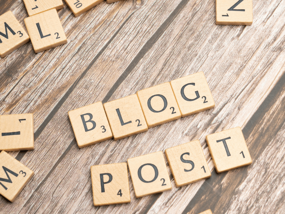 Letras de madeira formam as palavras “BLOG” e “POST” sobre uma mesa, destacando a criação de publicações para leitores.