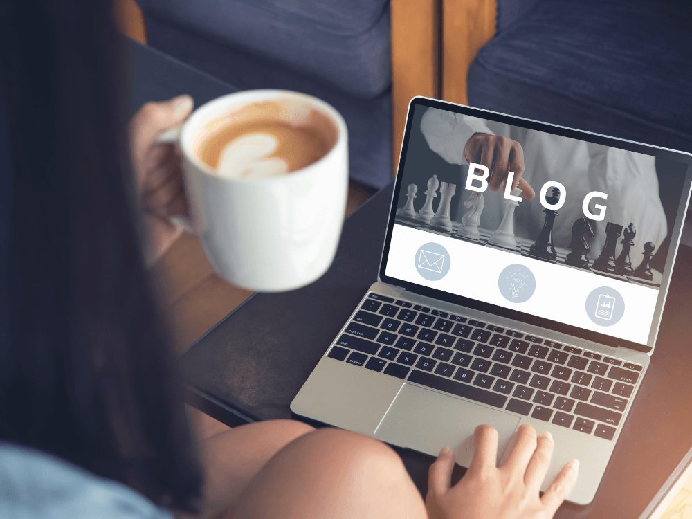 Pessoa utiliza notebook com uma página de blog aberta, enquanto toma café e acompanha o conteúdo produzido para publicação.